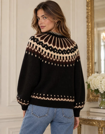 A woman wearing a black knit sweater with a cream and brown Fair Isle pattern around the yoke and cuffs, paired with blue jeans, standing in an elegant room with white walls and a gold-framed mirror.