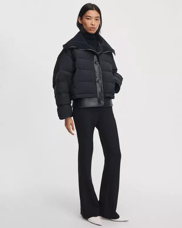 A model wearing a black cropped puffer jacket with a wide collar, black trousers, and white pointed shoes, standing against a light gray studio background.