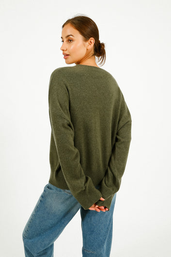 Woman wearing a green sweater and light-colored pants leaning against a wall.
