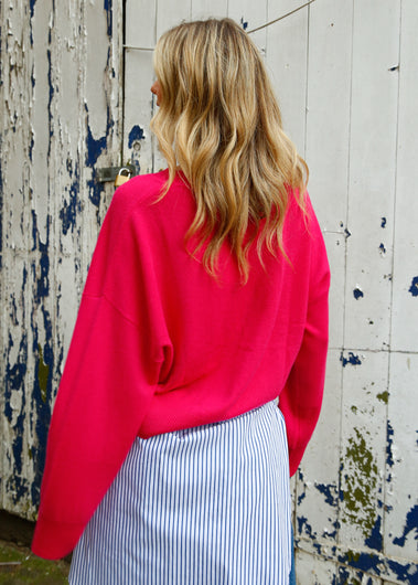 Woman wearing a bright pink sweater with a striped shirt tied around her waist, standing against a rustic wooden wall.