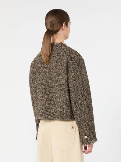 A woman modeling a cropped, long-sleeve tweed jacket in a brown and beige houndstooth pattern, paired with cream-colored trousers, against a plain white studio background.